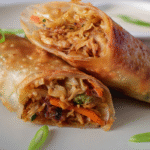 Crispy egg rolls cut in half, revealing a filling of shredded chicken, cabbage, carrots, and green onions on a white plate.