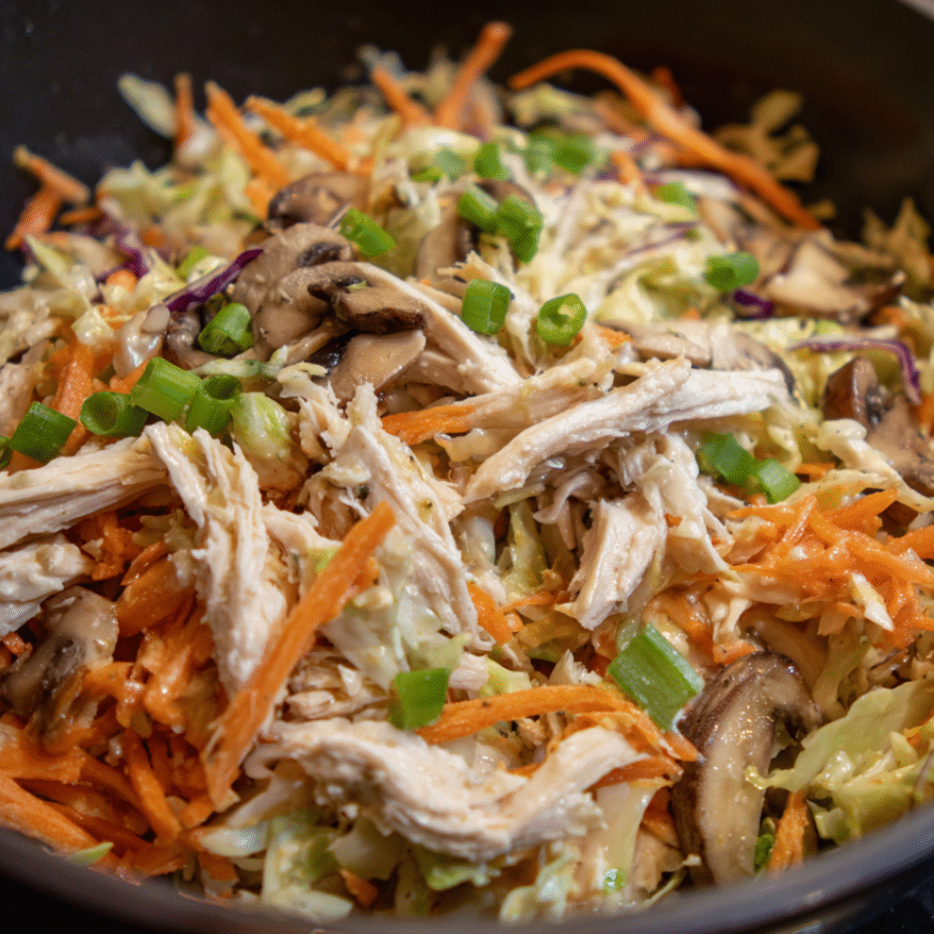 Close-up of shredded chicken, cabbage, carrots, mushrooms, and green onions being cooked in a pan as a filling for healthy egg rolls.